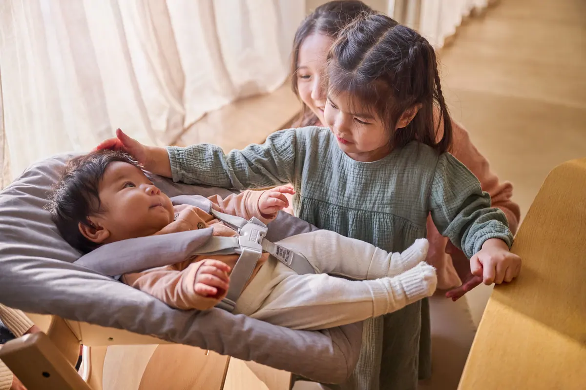 พี่สาวลูบหัวน้องแรกเกิดที่นอนอยู่ใน Tripp Trapp Newborn Set โดยมีคุณแม่อยู่ด้านหลัง