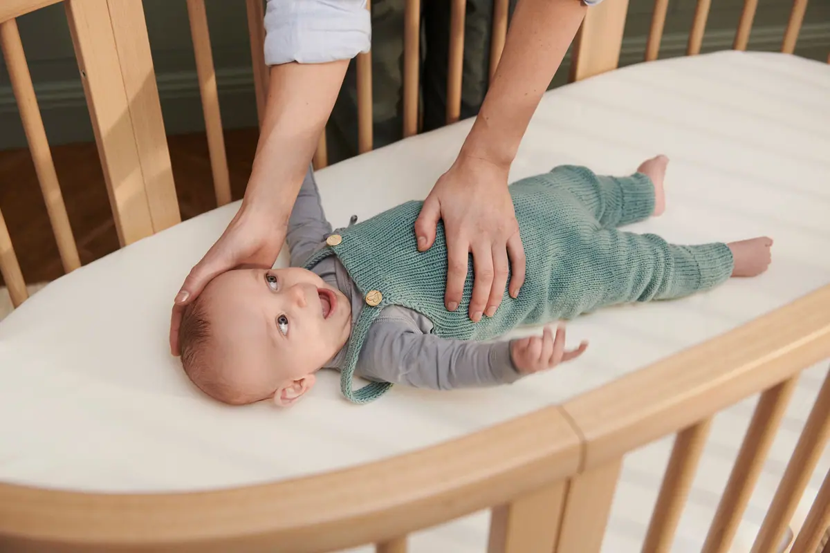 มือพ่อแม่วางบนตัวลูกที่ยิ้มอยู่ในเตียง Sleepi Bed