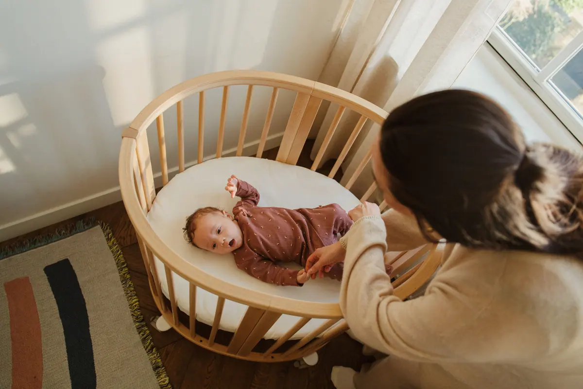 คุณแม่ก้มยิ้มให้ลูกที่นอนอยู่ในเตียง Stokke Sleepi Mini สีธรรมชาติข้างหน้าต่าง